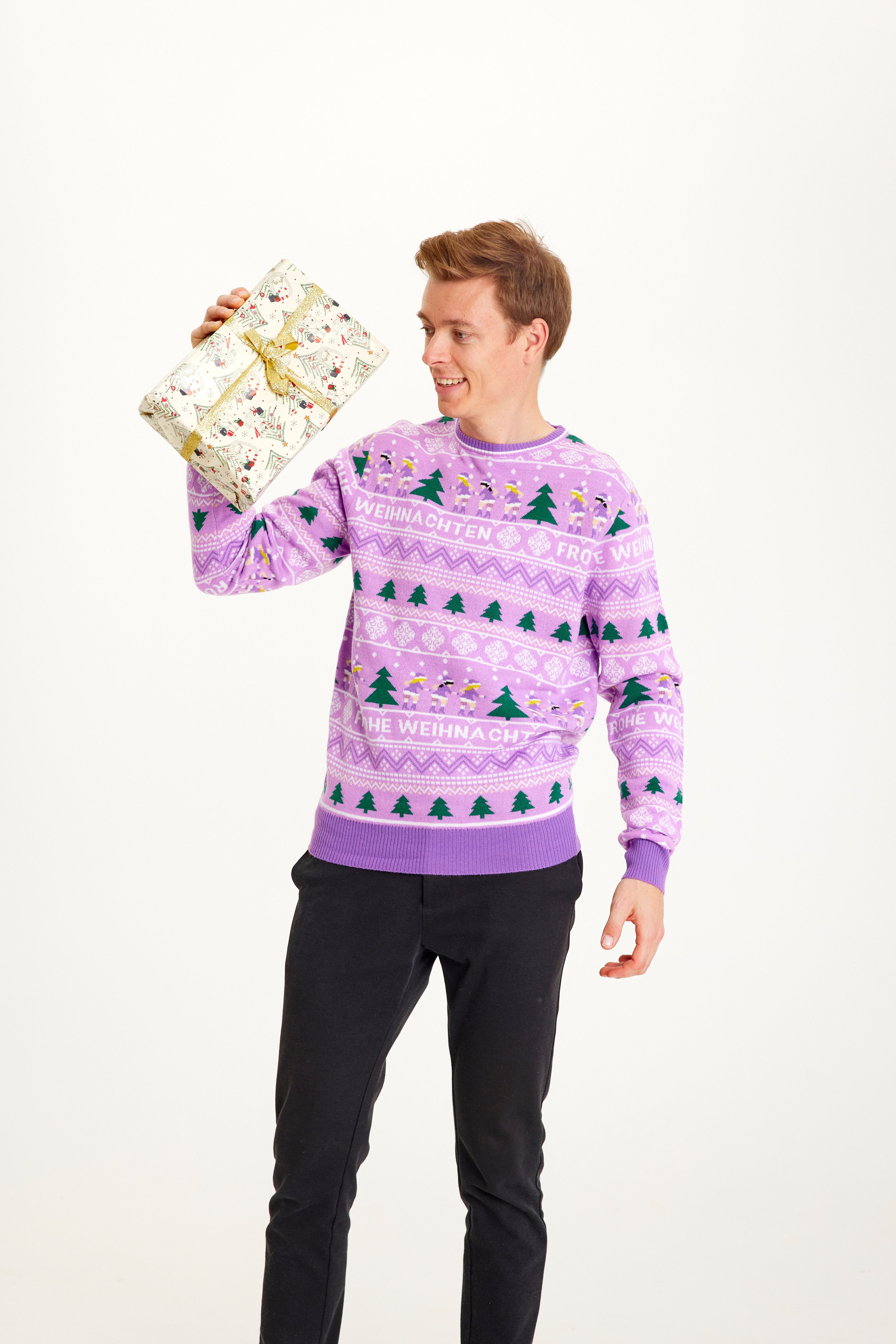 Een poserende man met een cadeau die een paarse kersttrui draagt met kerstbomen erop.